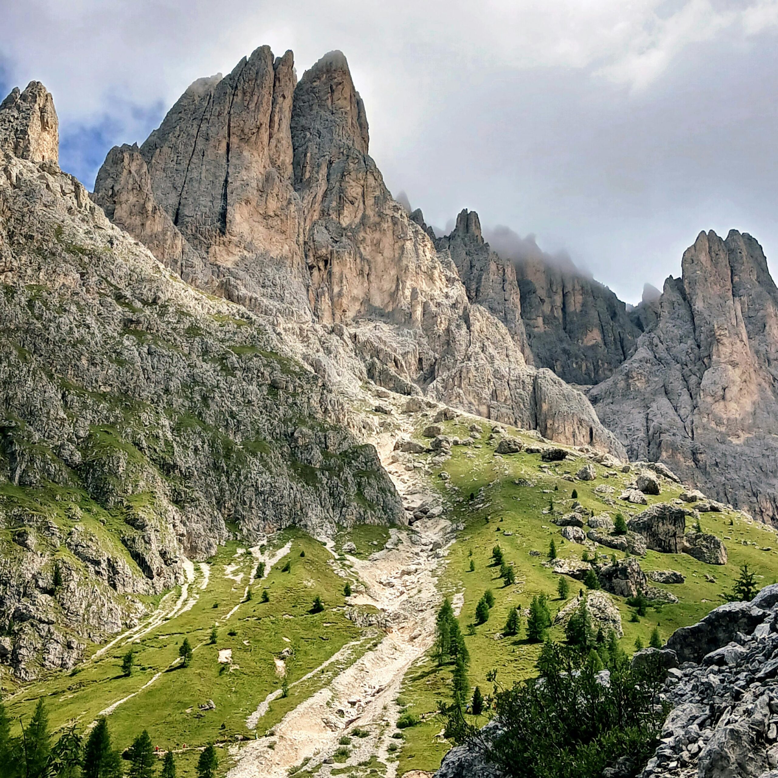 Breathtaking view of towering peaks in the Dolomites with lush green slopes and a rocky trail leading up to the clouds.